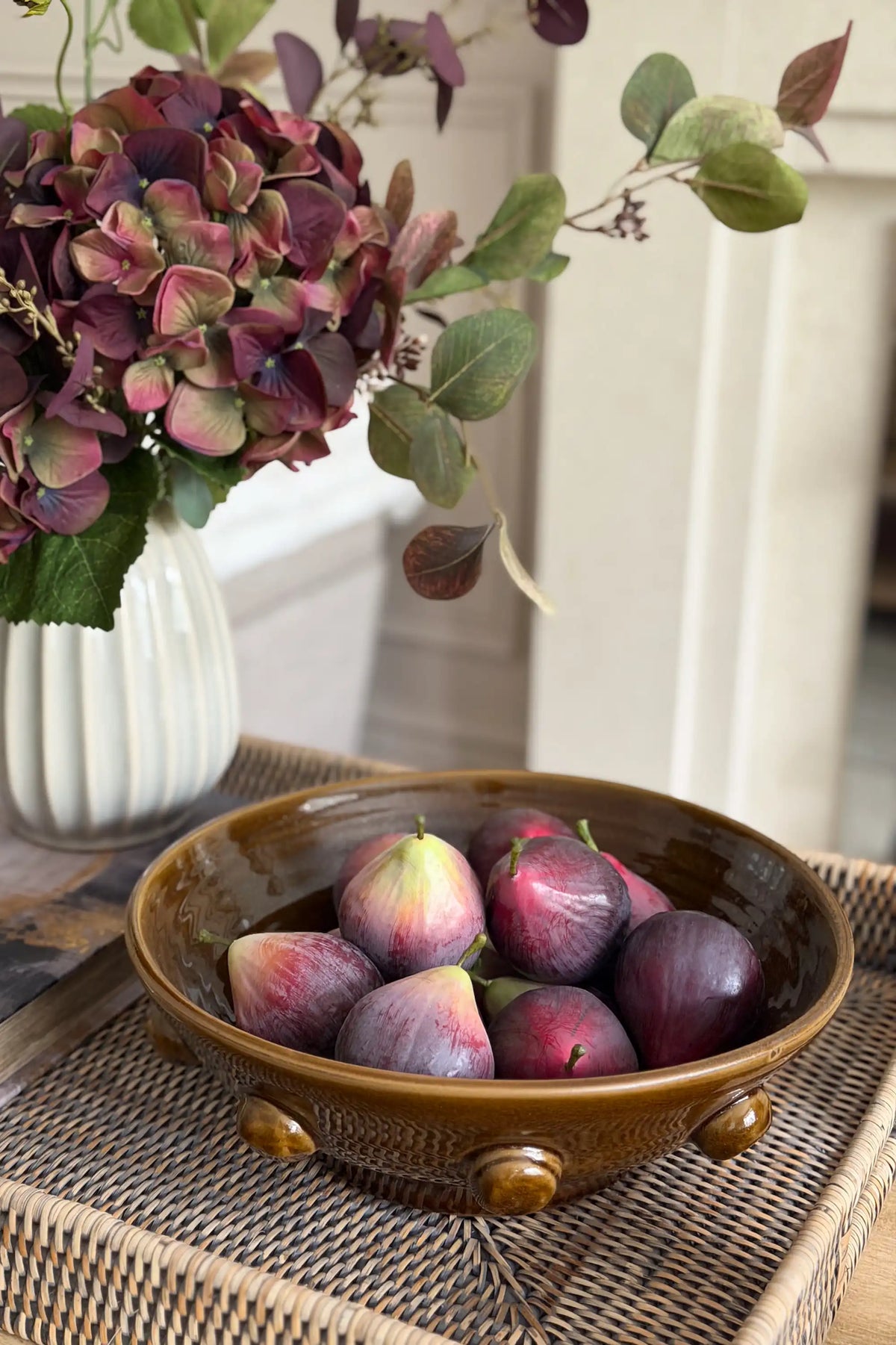 A ceramic bowl filled with fresh figs, styled on a woven tray next to a white vase with purple hydrangeas.